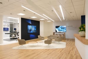 Modern interior of a brand space with wooden flooring, five beige armchairs around two round tables on a white rug, two large wall-mounted screens, the text ‘Passion For Life,’ and decorative plants on a counter.