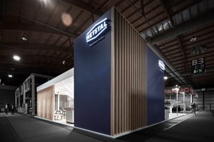 Ein moderner Messestand auf einer Fachmesse mit elegantem Design, vertikalen Holzlatten und einer großen blauen Wand mit dem Firmennamen „NETSTAL“ in weißer Schrift. Der Stand ist gut beleuchtet und enthält verschiedene Präsentations- und Sitzbereiche.