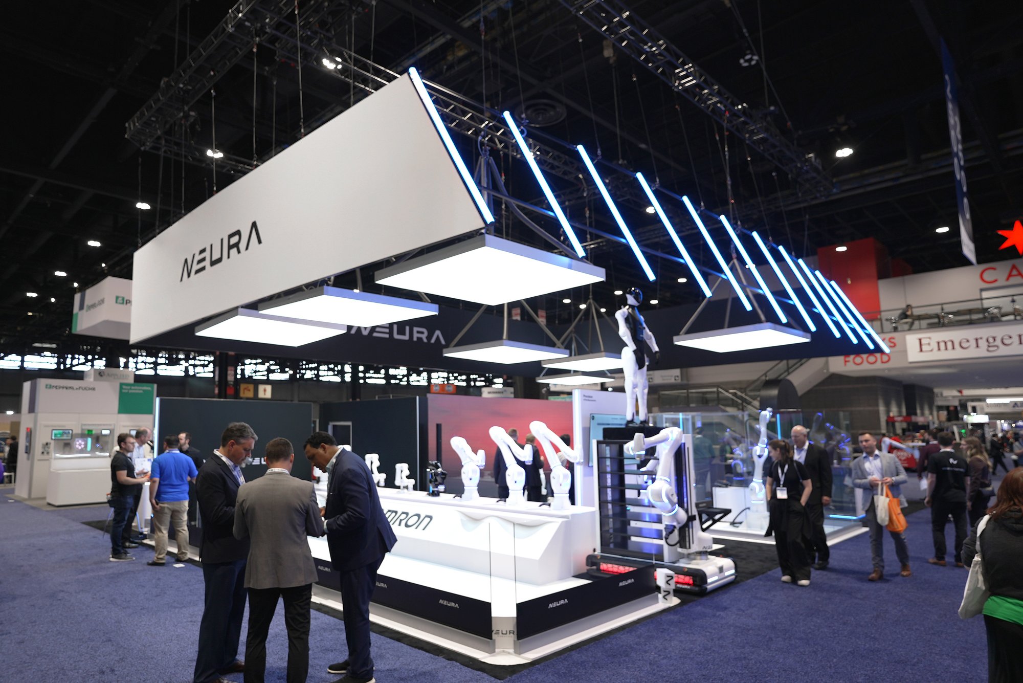 NEURA Robotics exhibition area at the trade fair with white banners and numerous visitors at the exhibition grounds. NEURA Robotics exhibition area at the trade fair with white banners and numerous visitors at the exhibition grounds.