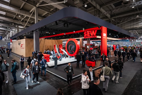 Stark besuchter SEW-EURODRIVE-Messestand mit hoch platziertem Logo in leuchtendem Rot, das über der Menschenmasse herausschaut..