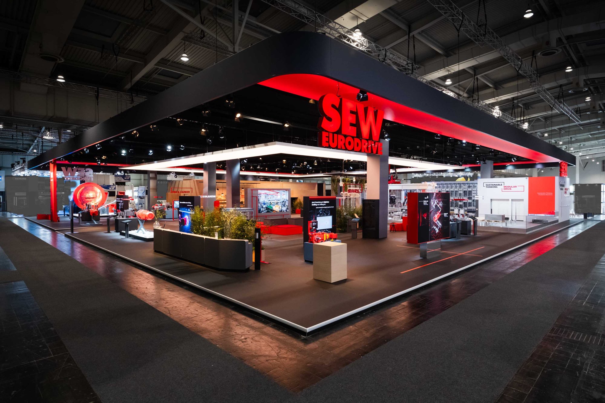 SEW-EURODRIVE exhibition stand at an industrial trade fair with large red structures and machine components, surrounded by visitors. SEW-EURODRIVE exhibition stand at an industrial trade fair with large red structures and machine components, surrounded by visitors.