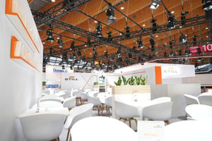 A view of a trade fair concept stand featuring numerous white chairs and tables, as well as stand walls cascading towards the rear.