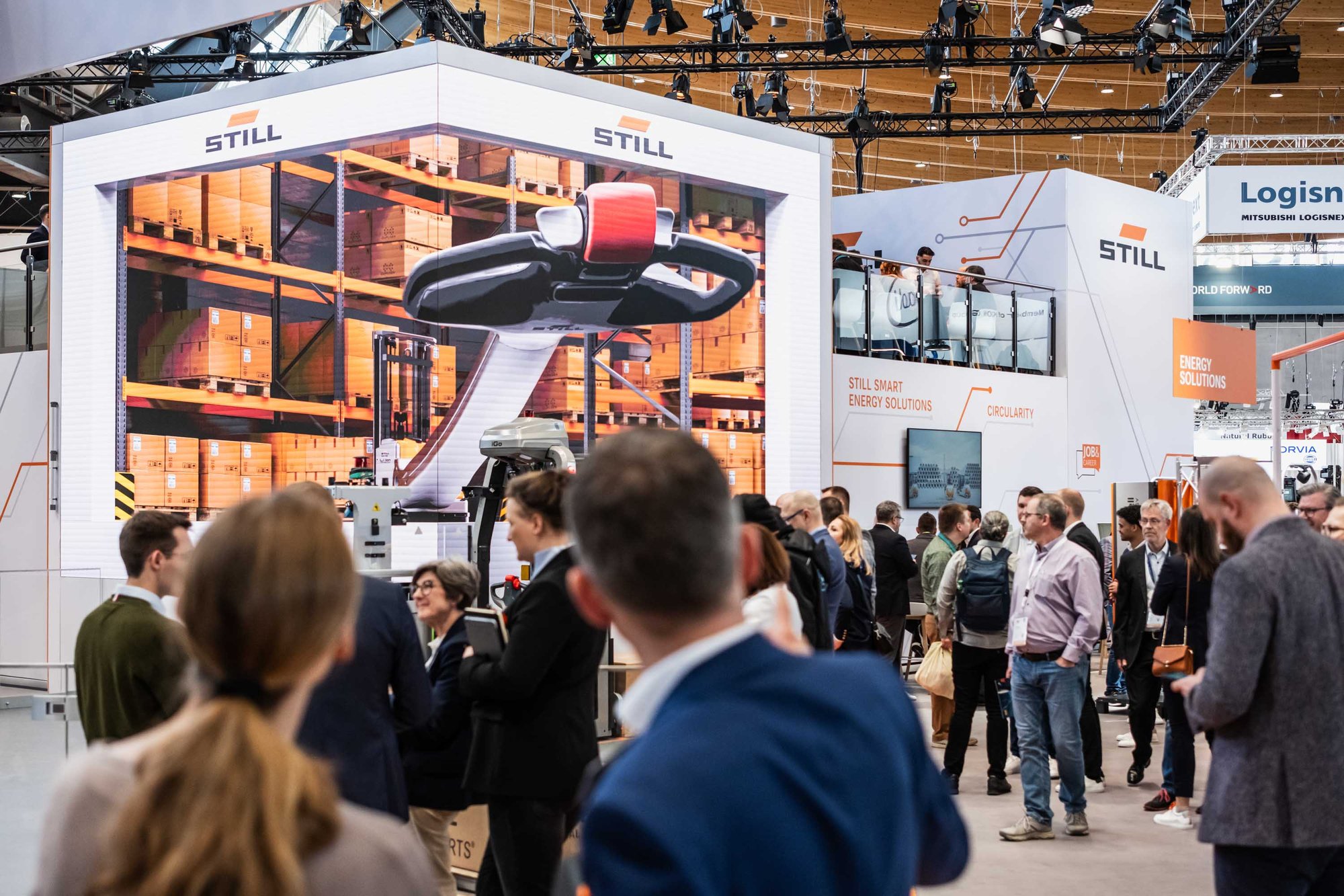 STILL exhibition stand at the logistics trade fair with automated storage solutions and robotics systems, visitors watching the presentation. STILL exhibition stand at the logistics trade fair with automated storage solutions and robotics systems, visitors watching the presentation.