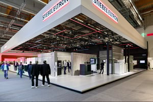 Eckansicht eines modernen Messestandes mit weißer Dachumrandung, Wänden im Betonstil und zahlreichen Besuchern auf der Fläche.