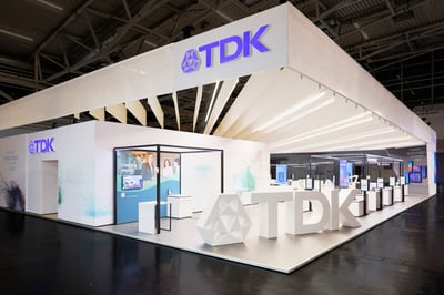 Heller und moderner Messestand von TDK Electronics mit hervorgehobenen blau leuchtenden Logo-Elementen und einem großen 3D-Druck des Logos am Eck des Standes.