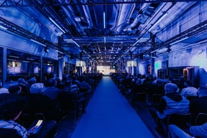 An event held in an industrial hall with atmospheric blue lighting and a large audience facing the main stage.