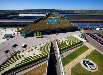 Aufsicht des Messe-Stuttgart-Geländes mit Grünflächen auf den Dächern, einem großen Vorplatz und Fahnen, die das Messe Stuttgart-Logo zeigen.