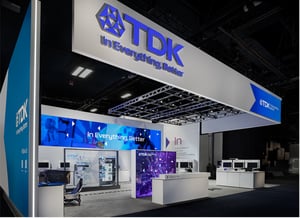 The TDK stand at CES in Las Vegas, featuring white and blue design elements, a large wall-mounted display and tables for visitors.