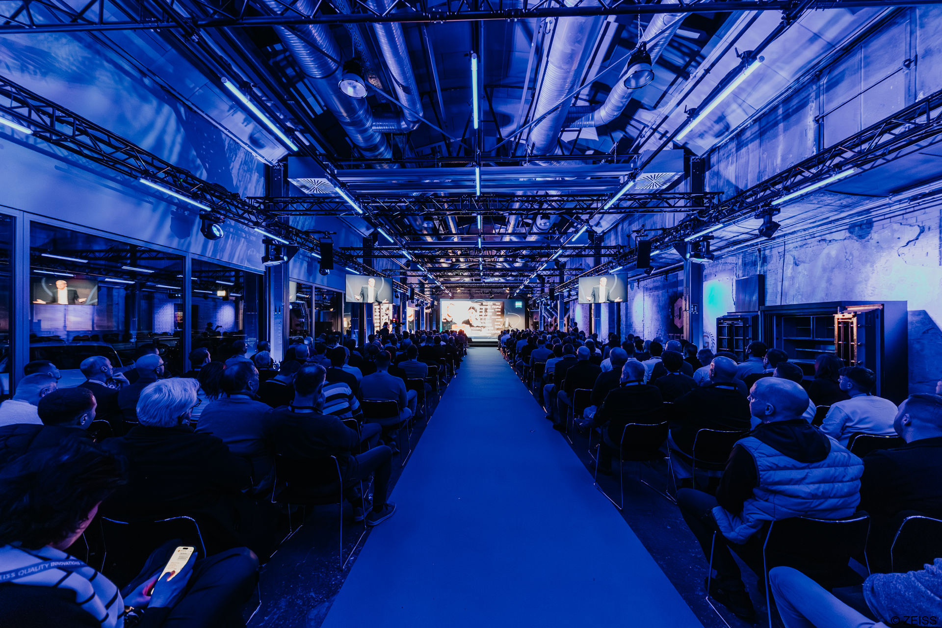 ZEISS conference in an industrial room with a big LED display on stage and lights surrounding sitting visitors that create an immersive digital experience.