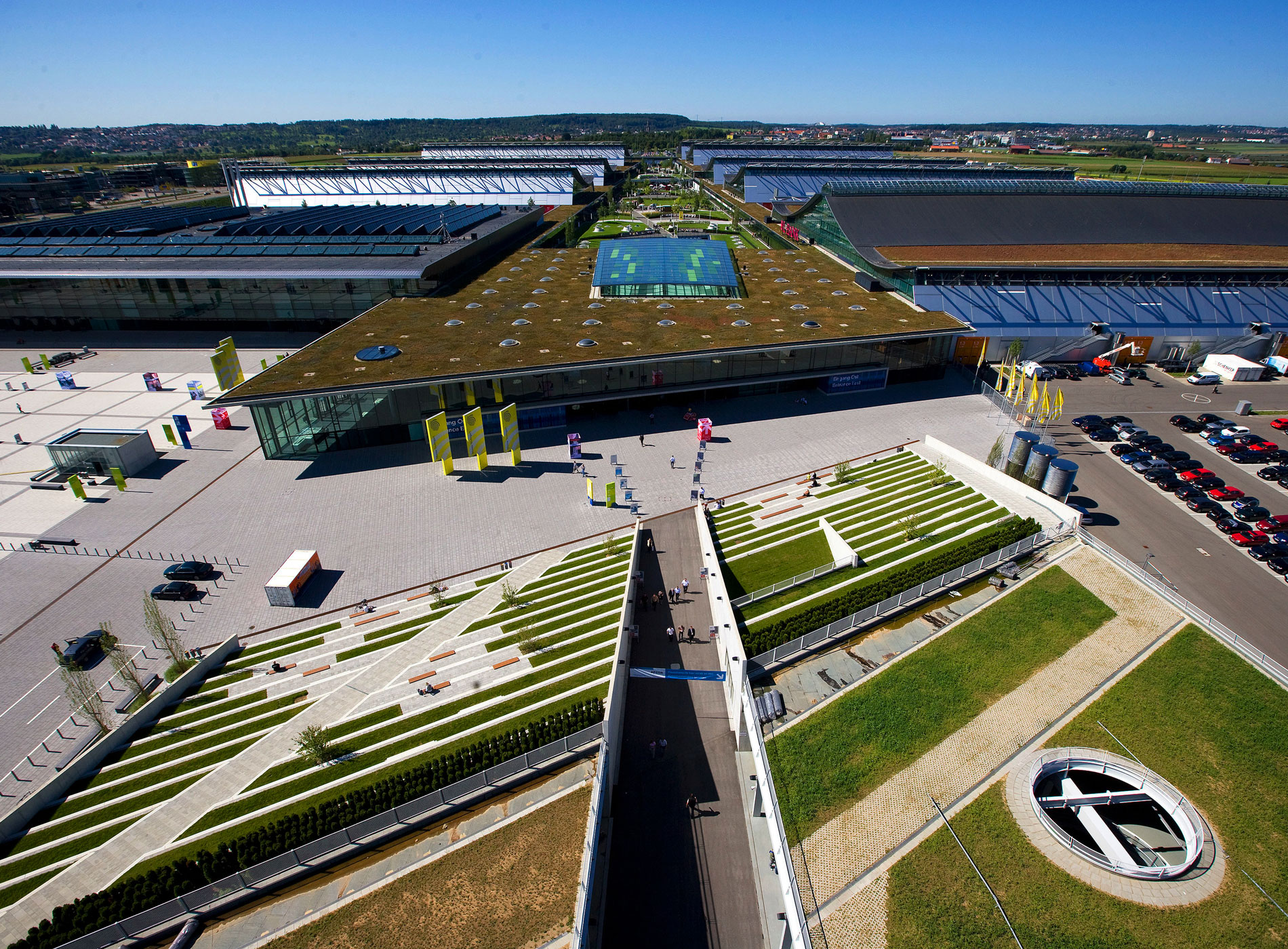 View of the Stuttgart Exhibition Center grounds with green spaces on the roofs, a large forecourt, and flags displaying the Stuttgart Exhibition Center logo.